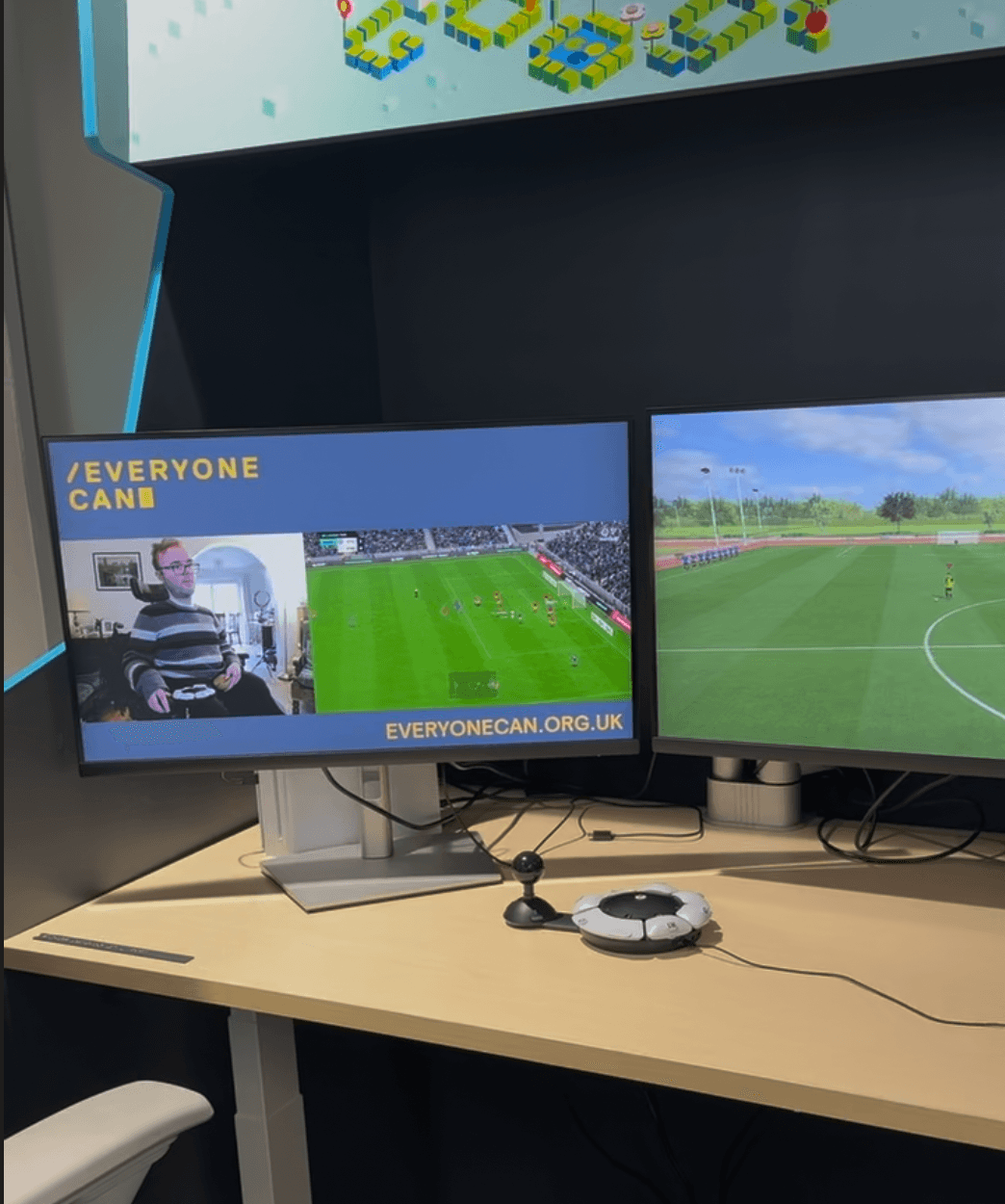An adaptive gaming setup at Google's Dublin Accessible Technology Lab, showing a switch-and-joystick controller on a desk in front of two monitors displaying an accessible sports game