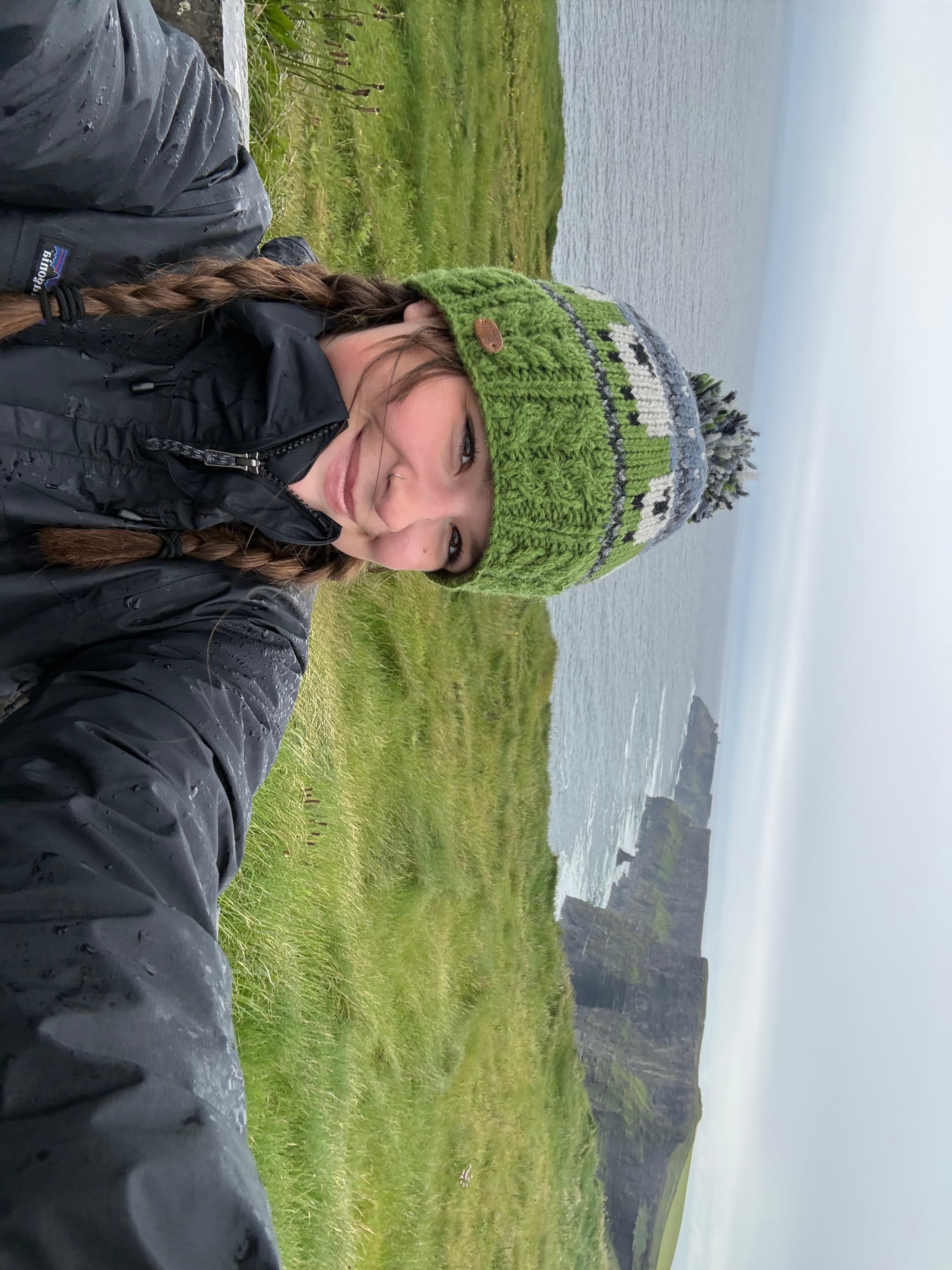 Mia smiling in a green knit hat and rain jacket at the Cliffs of Moher, with the cliffs and ocean behind her